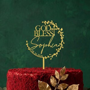 May include: A gold-coloured cake topper featuring the text GOD BLESS, the name Sophia, and the date 08.26.2026. The design includes a circular vine motif with leaves and a small cross integrated into the right side, supported by a vertical stem.