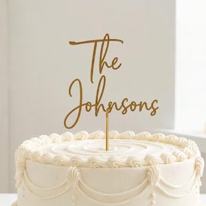 May include: A gold-colored cake topper with the words "The Johnsons" in elegant script. The topper is placed on top of a white frosted cake with decorative icing. The cake is a classic design, perfect for a wedding or anniversary.