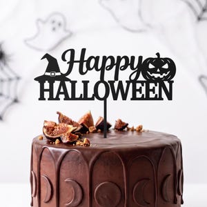 Może przedstawiać: Czarna ozdoba na tort z napisem Happy Halloween, kapeluszem czarownicy i dynią. Ozdoba jest umieszczona w okrągłym torcie czekoladowym z ciemną polewą i kawałkami świeżych fig na wierzchu.
