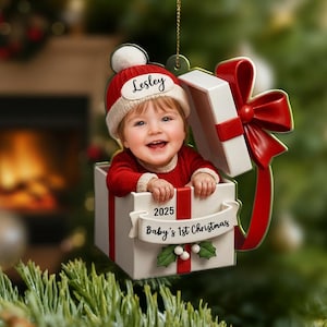 May include: Christmas ornament featuring a baby in a red outfit and hat, emerging from a white gift box with a red bow. The ornament has the text "Lesley", "2025", and "Baby's 1st Christmas".