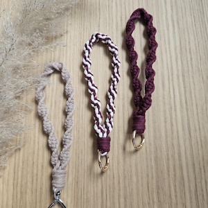 Puede incluir: Tres llaveros de pulsera de macramé con diseños de cordón trenzado. Uno es beige con un cierre plateado en forma de corazón, otro tiene un patrón en espiral blanco y burdeos con una anilla dorada, y otro es burdeos liso con una anilla dorada.