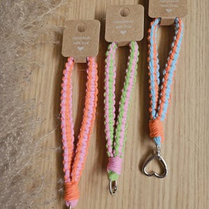 Pode incluir: Três cordões feitos à mão com fechos em forma de coração. Os cordões são feitos de cordão trançado em várias combinações de cores: rosa e laranja, verde e rosa e azul e laranja. Cada cordão tem uma pequena etiqueta que diz "Handmade with love."
