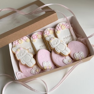 Peut inclure: Une boîte de biscuits de Pâques décorés. Les biscuits en forme de lapin et d'œuf sont décorés de glaçage blanc, de fleurs roses et jaunes, et de l'inscription "Happy Easter". La boîte est marron avec un couvercle transparent et un ruban rose.