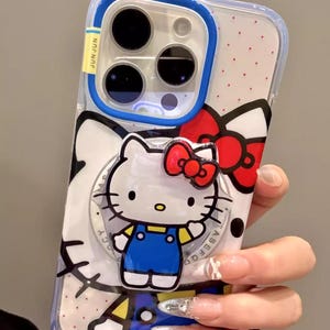 May include: Clear phone case featuring a Hello Kitty design. The case has a blue border and a pop-up ring holder. The Hello Kitty character is wearing a blue outfit and a red bow. The case has a polka dot pattern.
