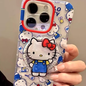 May include: Clear phone case featuring a repeating pattern of Hello Kitty characters in blue and white. The case has a red and blue border around the camera lenses and a Hello Kitty pop-up grip with a red bow. The case is designed for a smartphone.