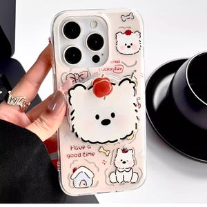 May include: Clear phone case with a light pink background and a cartoon dog design. The case features a 3D white dog with a red cherry on its head, along with other dog illustrations and the text "Have a good time".