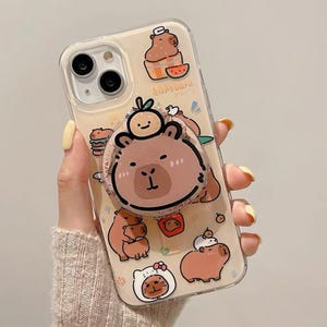 Puede incluir: Funda transparente para teléfono con temática de carpincho. La funda presenta ilustraciones de carpinchos de dibujos animados en varias poses, incluida una con una rodaja de sandía. Un agarre emergente con una cara de carpincho está adjunto.