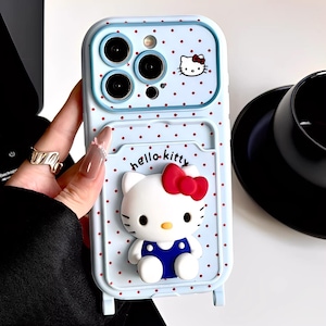 May include: Pale blue phone case featuring a Hello Kitty design. The case has a 3D Hello Kitty character with a red bow, a polka dot pattern, and the words "hello kitty". The case has a camera cutout and a card slot.