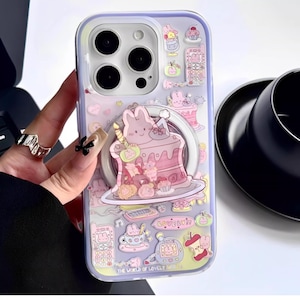 Könnte beinhalten: Transparente Handyhülle mit einem skurrilen Design mit einem rosa Kuchen mit einem Hasen, Cupcakes und anderen niedlichen Illustrationen. Die Hülle hat einen Ringhalter und die Worte "LOVELY" und "THE WORLD OF LOVELY PARTY".