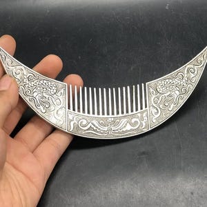 May include: A silver-colored comb with a crescent shape. The comb features intricate dragon and floral designs on the handle. The teeth of the comb are straight and evenly spaced.