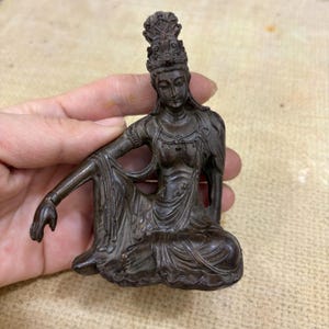 Puede incluir: Una estatua de color bronce de una figura sentada con un tocado adornado y túnicas fluidas. La figura está en una pose relajada, con un brazo apoyado en una rodilla doblada. La estatua tiene detalles intrincados y una apariencia desgastada.