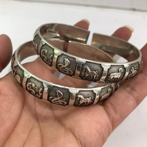 Può includere: Due braccialetti color argento con dettagliati disegni di animali. Ogni braccialetto presenta pannelli in rilievo con immagini di animali, tra cui serpenti e cavalli. I braccialetti sono tenuti in una mano, mostrando la loro intricata lavorazione.