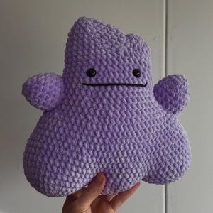 May include: A purple plush toy made of soft, textured yarn with a blob-like shape. It features two small rounded arms extending from the sides, two small black circular eyes, and a simple horizontal black line stitched to represent a mouth.