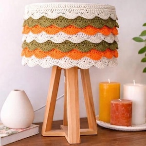 Peut inclure: Une lampe de table avec un pied en bois et un abat-jour cylindrique orné de couches de bordures crochetées en forme d'éventail, dans des tons crème, vert olive et orange vif. Le texte indique : PATRON DISPONIBLE en français et PATTERN AVAILABLE in english.