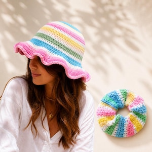 Op de afbeelding: Een gehaakte bucket hat met horizontale strepen in pastelroze, geel, groen en blauw met een golvende rand, vergezeld van een bijpassende gehaakte scrunchie met hetzelfde veelkleurige streeppatroon in een rond, geplooid ontwerp.