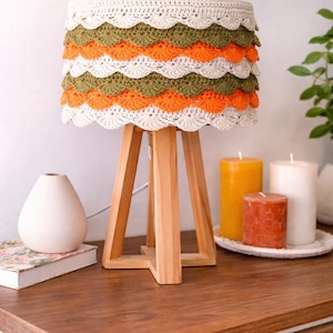 Peut inclure: Une lampe de table artisanale avec un pied en trépied en bois. L'abat-jour est crocheté avec un motif festonné en blanc, orange et vert olive. La lampe est posée sur une table en bois avec des bougies et un vase.