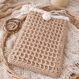 Peut inclure: Une pochette en crochet beige avec un motif texturé, une bordure blanche et une fermeture en pompon blanc. La pochette repose sur un plateau tressé avec un tissu en dentelle blanche. Une pile de bracelets de perles est à gauche.