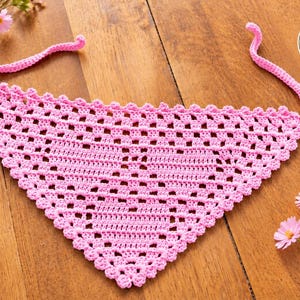 Può includere: Una bandana triangolare all'uncinetto di colore rosa con un motivo a griglia traforata, un bordo smerlato e due lunghi lacci. Un badge bianco circolare nell'angolo in alto a destra mostra la bandiera francese e il testo PATRON EN français.