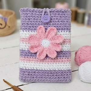 Puede incluir: Una funda de teléfono de ganchillo hecha a mano en tonos morado, blanco y rosa. La funda presenta una gran flor rosa con un centro blanco y un cierre de botón. La funda mide aproximadamente 15 cm de alto y 9 cm de ancho.