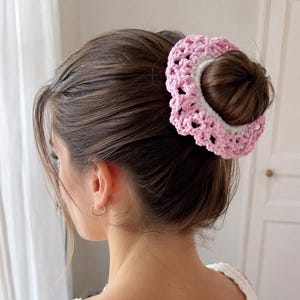 May include: A pink and white crocheted hair accessory, worn in a bun. The accessory is a circular design with a scalloped edge. The hair is brown and styled in a low bun. The person is wearing gold hoop earrings.
