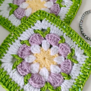Peut inclure: Deux carrés en crochet avec des motifs floraux. Chaque pièce présente une fleur jaune centrale entourée de pétales blancs et d'accents lavande. Les bords sont bordés de fil vert vif.