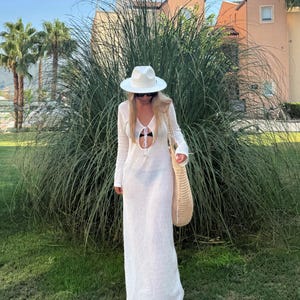 May include: A white, long-sleeved, floor-length dress with a keyhole cutout. The person is wearing a white hat, sunglasses, and carrying a woven tote bag. The background includes green grass, tall plants, and buildings.