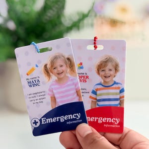 Op de afbeelding: Twee noodinformatiekaarten worden in een hand gehouden. Eén kaart heeft een foto van een meisje en de tekst "My name is MAYA WINC" en "I am anaphylactic to nuts. I always carry an epipen in my bag." De andere kaart heeft een foto van een jongen.