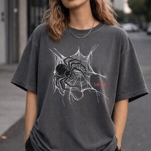 A woman wearing a dark gray t-shirt with a white spider web and black spider graphic, and the words "STAY AWAY" in red, paired with a black beanie.