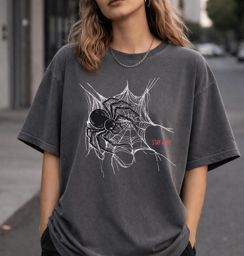 A woman wearing a dark gray t-shirt with a white spider web and black spider graphic, and the words "STAY AWAY" in red, paired with a black beanie.