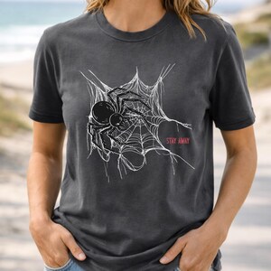 A woman in a dark gray t-shirt featuring a white spider web and black spider graphic, with "STAY AWAY" in red text, standing on a beach path.
