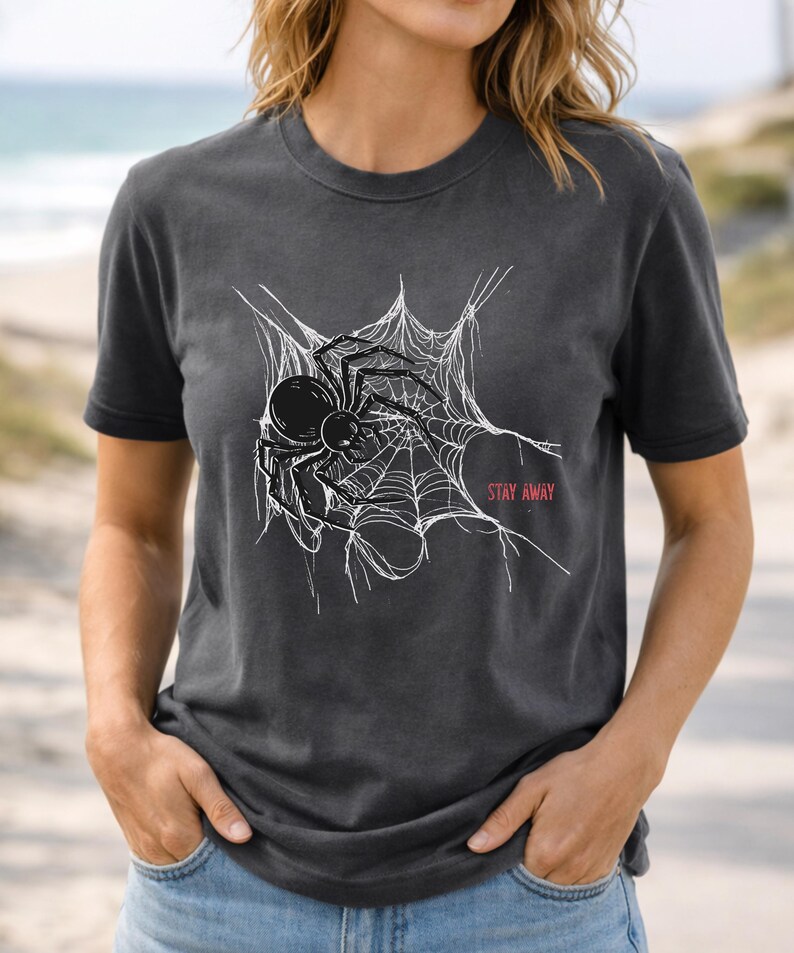 A woman in a dark gray t-shirt featuring a white spider web and black spider graphic, with "STAY AWAY" in red text, standing on a beach path.