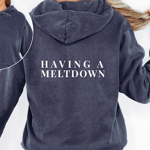 A person wearing a dark blue vintage-wash hoodie featuring the text HAVING A MELTDOWN in white capital letters across the back, with an inset showing the front.