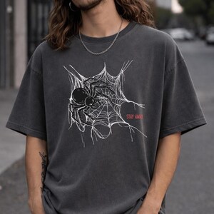 A man with long hair wearing a dark gray t-shirt with a white spider web and black spider graphic on the chest, with "STAY AWAY" in red text.