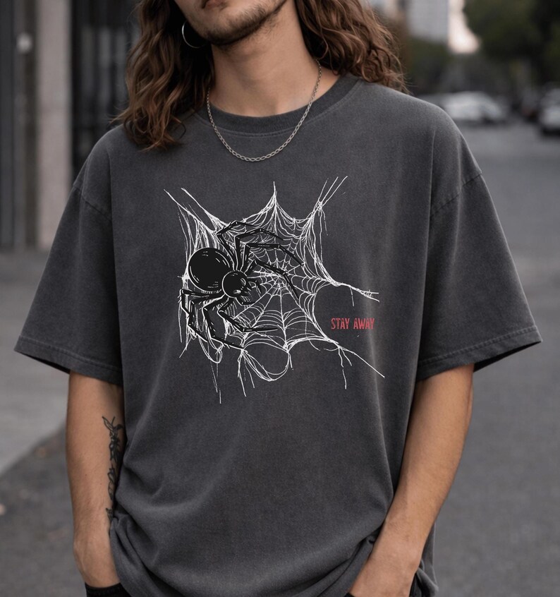 A man with long hair wearing a dark gray t-shirt with a white spider web and black spider graphic on the chest, with "STAY AWAY" in red text.