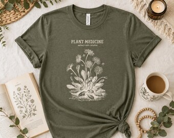 May include: Olive green t-shirt with the words "PLANT MEDICINE" and a botanical illustration. The shirt also has the words "ancient • wild • intuitive" printed on it. A book, a cup of coffee, and a candle are in the background.