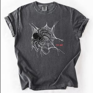 A dark gray t-shirt with a white spider web and black spider graphic, and the words "STAY AWAY" in red, laid flat on a white surface.