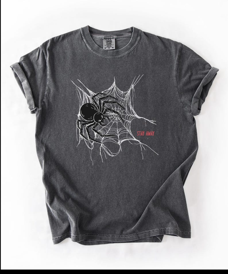 A dark gray t-shirt with a white spider web and black spider graphic, and the words "STAY AWAY" in red, laid flat on a white surface.