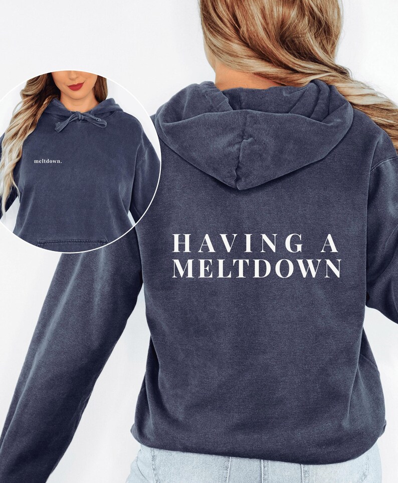 A person modeling the dark blue hoodie from behind with the text HAVING A MELTDOWN on the back, and an inset showing the front text reading meltdown in lowercase.