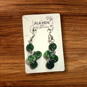 May include: A pair of dangling earrings featuring clusters of green buttons. The earrings are attached to a white card with the text "ALA MEN ART COLLECTION" and a hand-drawn illustration. The earrings hang against a wooden background.