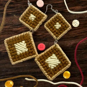 May include: Handmade square earrings with a brown and cream woven design. The earrings have a cream-colored diamond pattern in the center and are accented with a gold border. The earrings are displayed with colorful buttons and yarn.