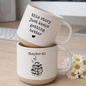 May include: Two stacked white ceramic mugs with a beige base. The top mug reads "this story just keeps getting better" with a heart. The bottom mug says "Chapter 60" with a stack of books and a teacup design.