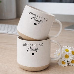 Può includere: Due tazze in ceramica bianca impilate. Ogni tazza ha una base beige e una sezione superiore bianca con manico. Il testo "chapter 40 Charlotte" e un cuore nero sono stampati sulla parte anteriore di ogni tazza.