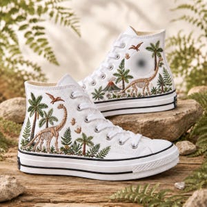 Puede incluir: Un par de zapatillas altas de lona blanca con bordados detallados de esqueletos de dinosaurios prehistóricos, palmeras, helechos y un volcán. Los zapatos tienen cordones blancos, puntera de goma y una raya negra en la suela de goma.