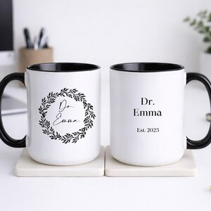 May include: Two white ceramic mugs with black handles and interior. One mug features a wreath design with the text "Dr. Emma". The other mug has the text "Dr. Emma" and "Est. 2025". Both mugs are on square coasters.