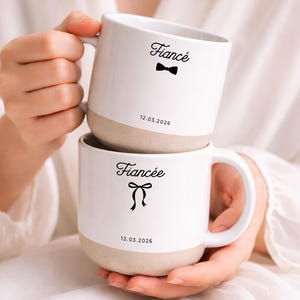 Może przedstawiać: Dwa białe ceramiczne kubki, jeden na drugim. Na górnym kubku napis "Fiancé" z muchą, a na dolnym "Fiancée" ze wstążką. Oba kubki mają nadrukowaną datę "12.03.2026". Kubki są trzymane przez osobę.