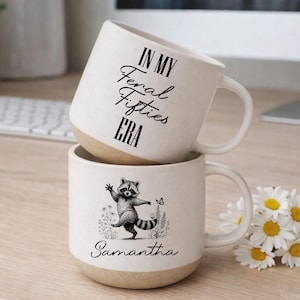 Puede incluir: Dos tazas de cerámica blanca apiladas. La taza superior tiene el texto "IN MY Feral Fifties ERA". La taza inferior presenta una ilustración de mapache y el nombre "Samantha". Ambas tazas tienen una base beige.