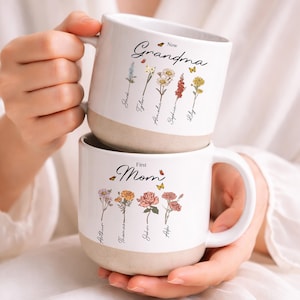 May include: Two white ceramic mugs with floral designs and the words "Now Grandma" and "First Mom". The mugs have a beige base and feature names and butterflies. A person is holding the mugs.