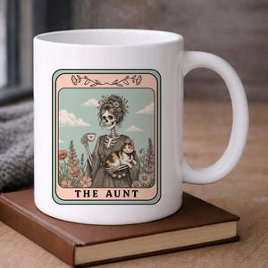 May include: White ceramic mug featuring a whimsical illustration of a skeleton holding a cat and a cup of tea, set against a floral and sky background. The text "THE AUNT" is displayed at the bottom of the image.
