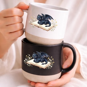 May include: Two ceramic mugs, one white and one black, each featuring a whimsical illustration of a dragon resting on an open book. The dragon is dark blue with starry accents and wings. The mugs have a beige base.