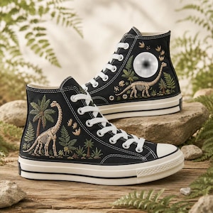 Puede incluir: Zapatillas altas de lona negra con cordones blancos y punteras de goma. Los laterales están decorados con un bordado detallado de un esqueleto de dinosaurio, palmeras, helechos y pequeñas huellas en tonos verde, marrón y crema.